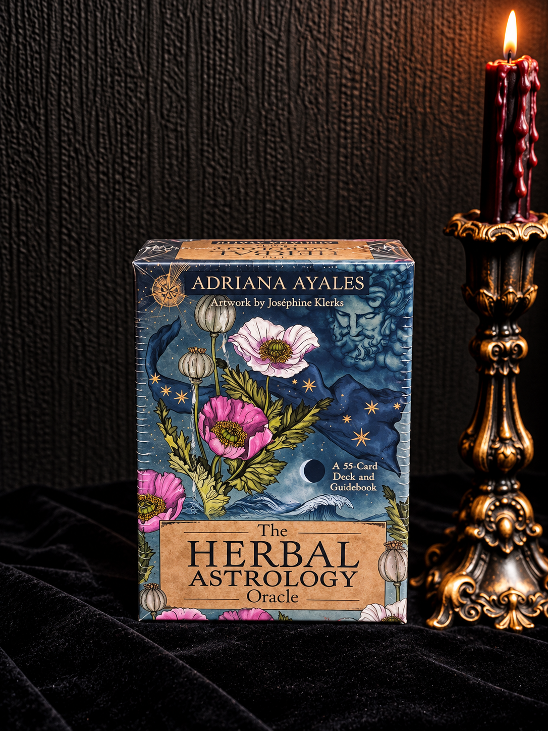 The Herbal Astrology Oracle card deck box by Adriana Ayales, styled on a black velvet surface next to a lit dark red wax candle in an ornate gold holder.