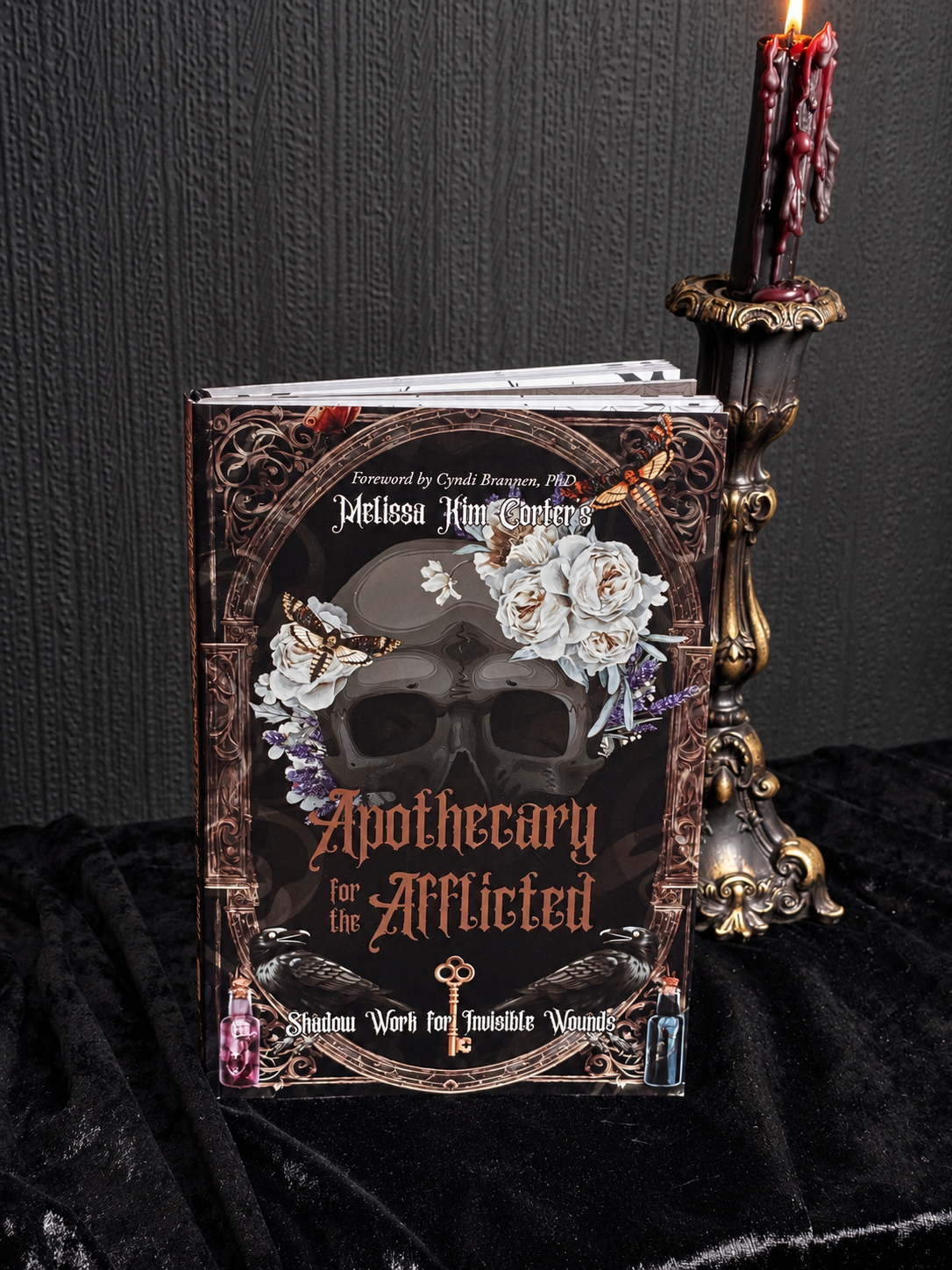 Apothecary for the Afflicted by Melissa Kim Corter, dark illustrated book cover featuring a skull, white roses, moths, ravens and ornate apothecary imagery.