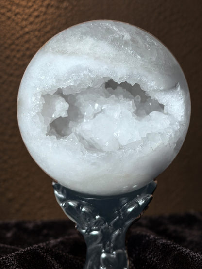 A polished white crystal geode sphere with a hollowed-out crystalline interior, displayed on a dark ornate stand against a brown textured background.