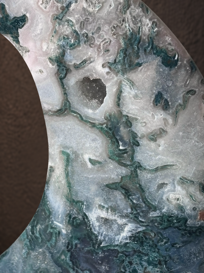 A close-up of a crescent-shaped stone or agate slice with swirling patterns of teal green, white, and gray, set against a dark brown textured background.