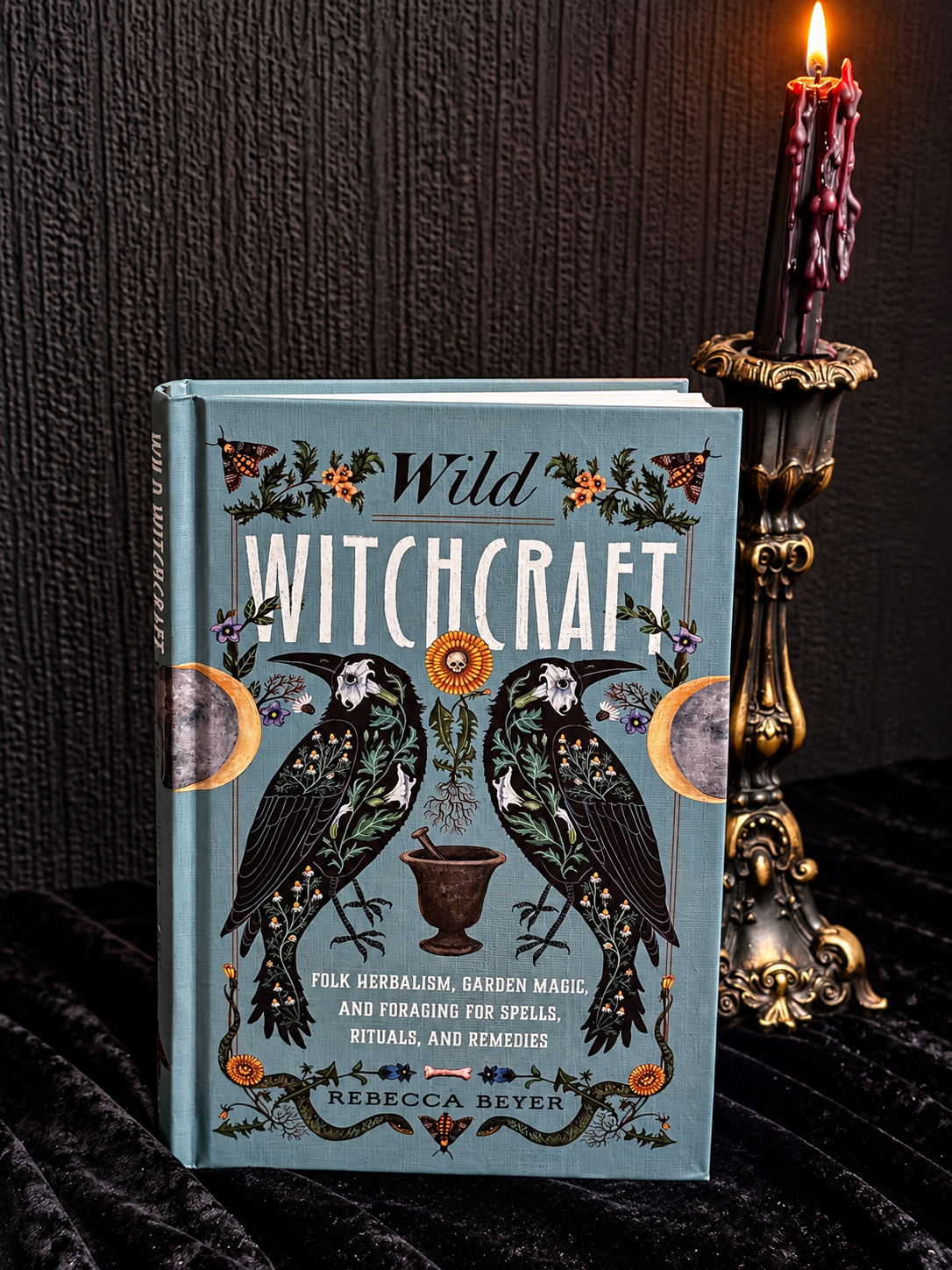 Wild Witchcraft by Rebecca Beyer, blue-green hardcover book featuring two black birds, crescent moons, botanical illustrations and flowers.