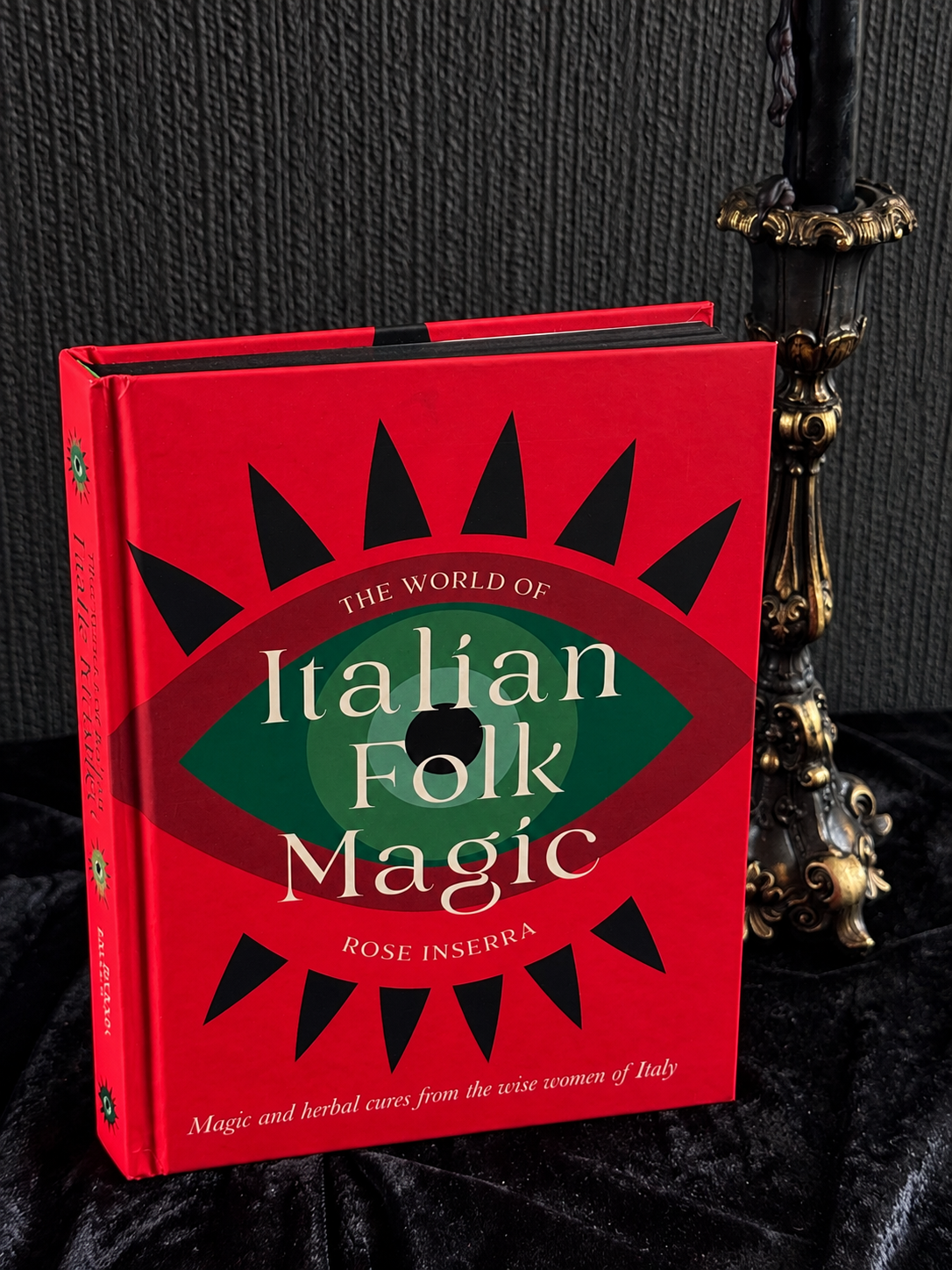 The World of Italian Folk Magic by Rose Inserra, vivid red hardback book featuring a large green eye motif with black radiating shapes and cream lettering.